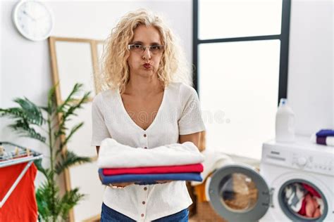Middle Age Blonde Woman Holding Folded Laundry After Ironing Puffing Cheeks With Funny Face