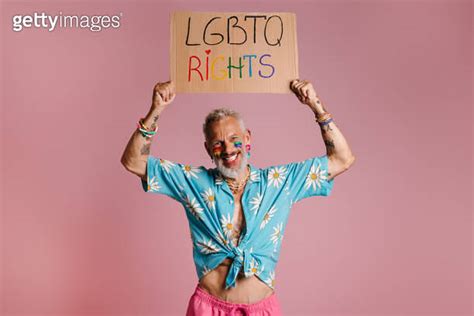 Happy mature gay man with rainbow flag make up holding banner against pink background 이미지