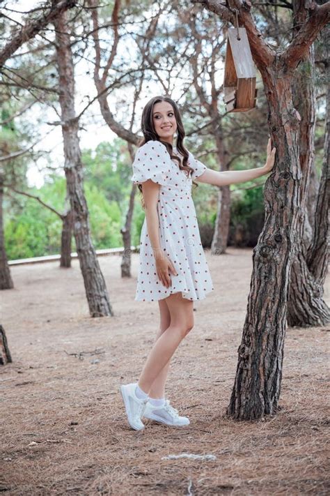 Beautiful Slim Brunette Woman In Summer White Dress Walking Among Trees In Park Stock Photo