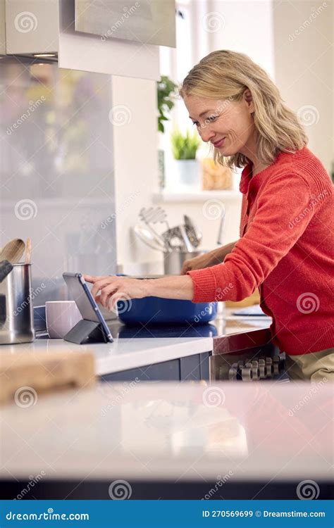 Mature Woman In Kitchen At Home Cooking Meal Following Recipe On Digital Tablet Stock Image