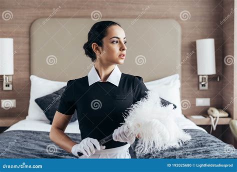 Brunette Maid In Apron And Gloves Holding Duster And Looking Away In Hotel Room Stock Photo