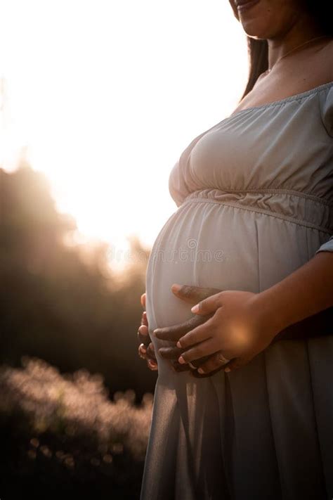 Pregnant Latina Woman In A Dress Standing In Lush Green Sunlit Park Hands Gently Cradling Her