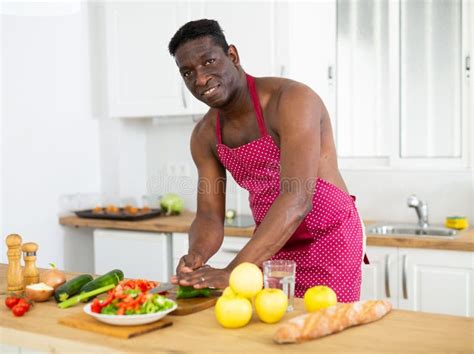 Man From Africa With A Naked Torso In An Apron Prepares A Vegetable Salad In The Kitchen Stock