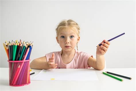 Fille Enfant Blonde Aux Yeux Bleus Dessine Avec Des Crayons De Couleur Assis Une Table Blanche
