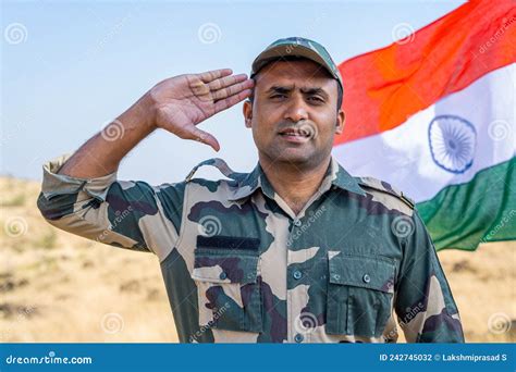 Army Officer Saluting By Looking At Camera In Front Of Waving Indian