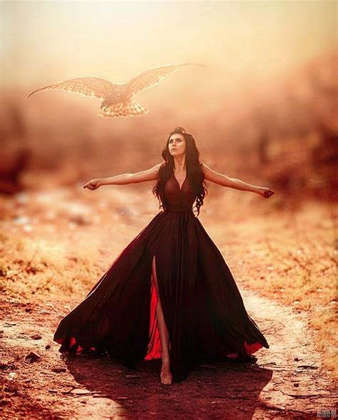 A Woman In A Long Black Dress Standing On A Dirt Road With An Owl