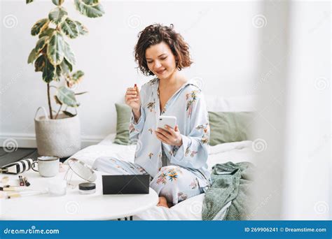 Junge Brunette Frau In Blauen Pyjamas Schminkt Sich Mit Handy Sitzend Im Bett Zu Hause Stockbild