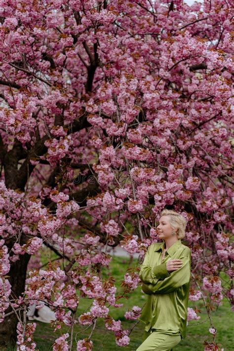 A Blonde Woman In Green Outfit Poses Surrounded By Cherry Blossoms Outdoors Stock Photo Image
