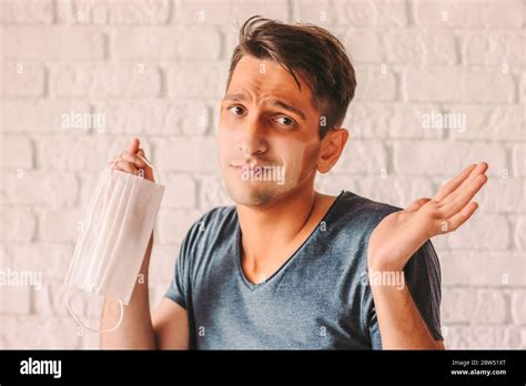 Portrait of confused man with funny sun tan lines on face holding protective mask in hand. Young ...