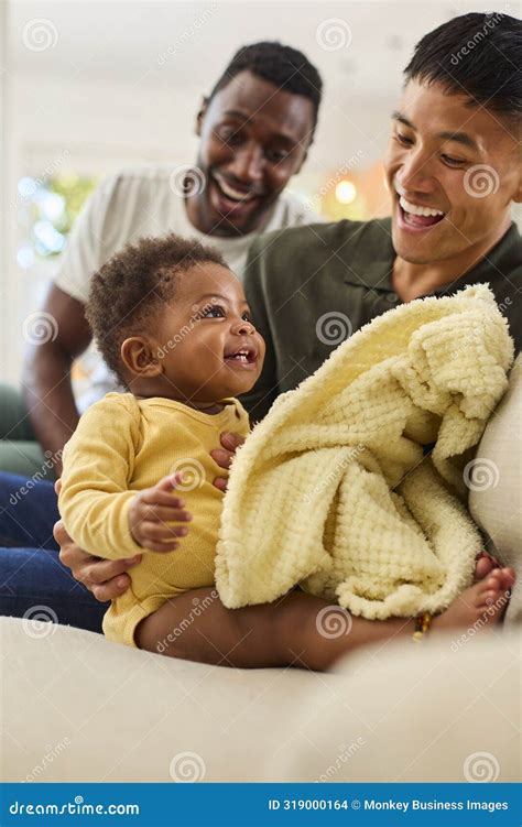 Same Sex Male Couple Or Friends Playing With Baby Sitting On Sofa At Home With Blanket Together
