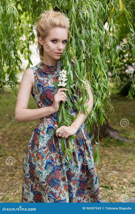 Beautiful Sweet Tender Girl Blonde With Blue Eyes Stands Near A Tree With Long Branches With A