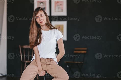 Beautiful Brunette Girl With Long Flowing Hair Dressed In White T Shirt And Jeans Poses Sitting