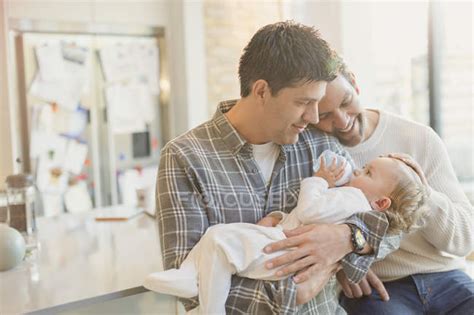 Affectionate Male Gay Parents Holding Baby Son With Bottle Kitchen Baby Bottle Stock Photo