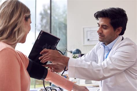 Male Doctor Or GP Examining Mature Female Patient Taking Blood Pressure With Sphygmomanometer