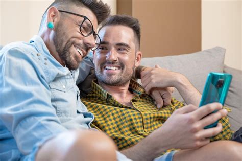 Alegre casal gay sentado no sofá abraçando e olhando para o celular assistindo a um filme
