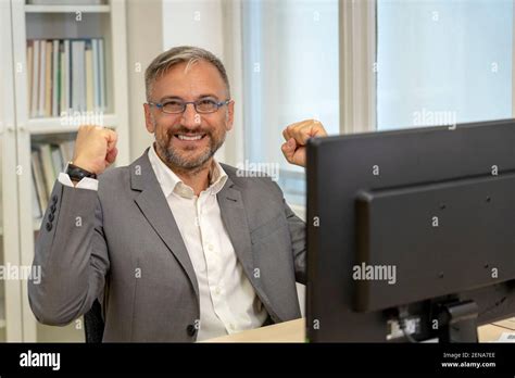 Successful Mature Businessman Sitting At Office Desk Raising Hand In