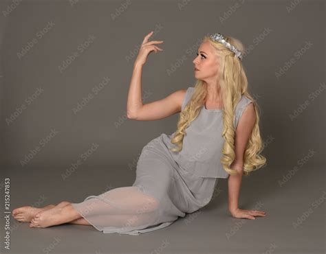 Full Length Portrait Of Blonde Woman Wearing Crown And Pale Blue Dress Seated Pose Against A