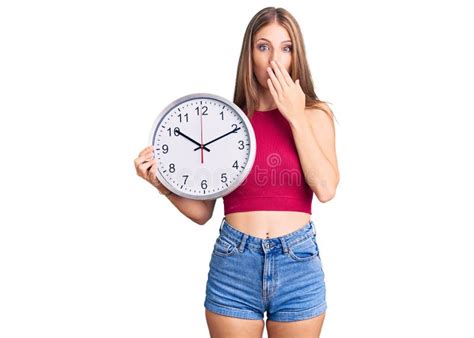 Jeune Belle Femme Blonde Tenant Grande Horloge Couvrant La Bouche Avec La Main Choquée Et Peur