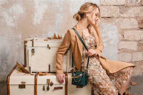 Jolie femme blonde élégante en manteau beige assis sur des valises contre le mur dans la rue
