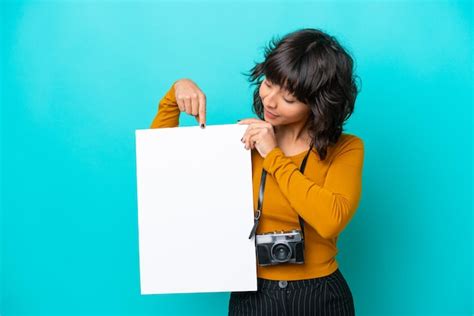 Jovem fotógrafa latina isolada em fundo azul segurando um cartaz vazio expressão feliz e