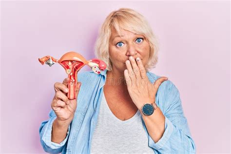 Middle Age Blonde Woman Holding Anatomical Model Of Female Genital Organ Covering Mouth With