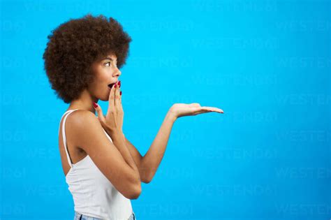 Latina Frau mit Afro Haar überrascht Hand ausgestreckt zeigt Studio Bild mit blauem Hintergrund