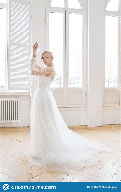 Beautiful Slender Blonde In The Evening Sun In A Long White Dress Near A Large Window Perfect