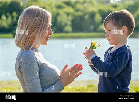 Cute Baby Son Gives A Gift Flowers To His Beautiful Blonde Mother In The Park On A Background