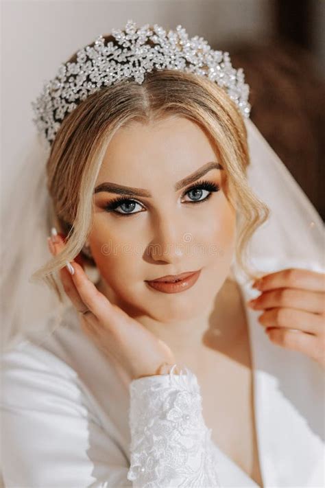 Wedding Portrait Blonde Bride In A Silk Suit And Tiara Poses Behind A Sheer Veil While Sitting