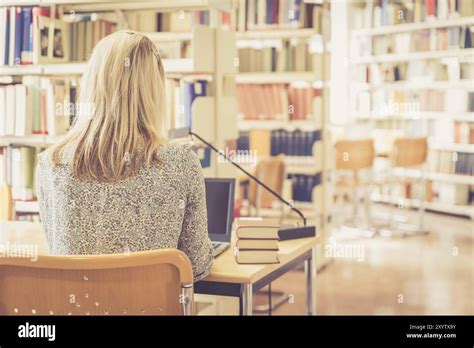 Blonde Female Student Is Sitting At The Desk With Pile Of Books University Library Stock Photo