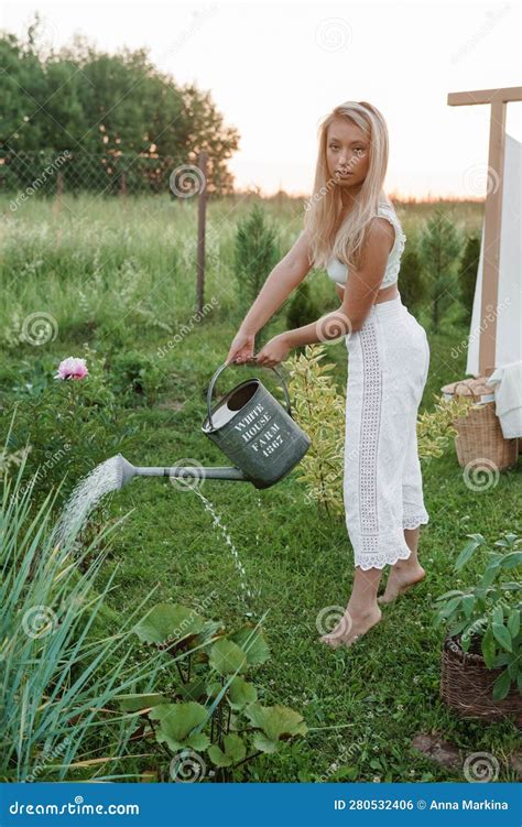A Blonde Woman Is Watering Flowers In The Garden In The Courtyard Of A Village House Stock