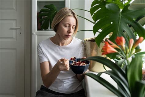 Mature Blonde Woman Eats Breakfast In Bowl Sitting Near Windowsill With Flowerpots Stock Image