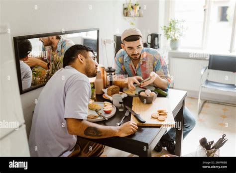 Gay Couple Sharing Smart Phone With Each Other While Having Food On Dining Table At Home Stock