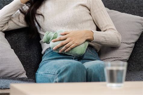 Woman Using Hot Water Bottle For Period Pain Relief On Sofa Sick At Home Concept Stock Photo