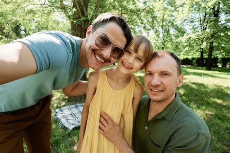 Retrato de feliz pareja masculina gay abrazando con su hija adoptiva y sonriendo a la cámara al