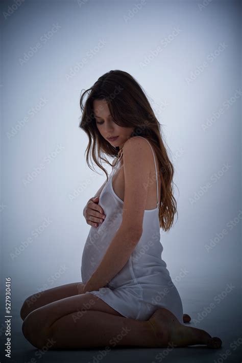 Beautiful Pregnant Brunette Hair Woman In Elegant White Dress Posing To Photographer In Studio