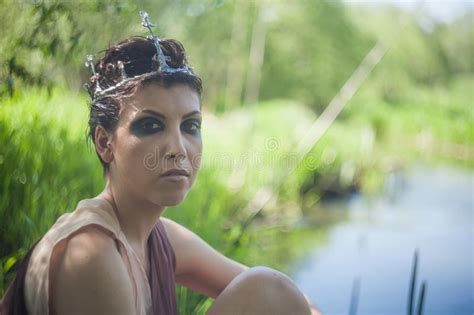 Slender Brunette In A Brown Dress With A Crown On Her Head Sits On The Shore Of A Forest Pond