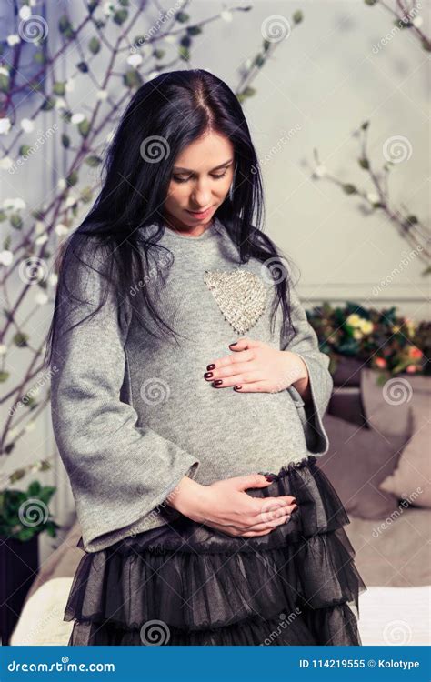 Attractive Pregnant Brunette Woman Keeping Hands On Belly Standing Near Bed In The Bedroom Stock
