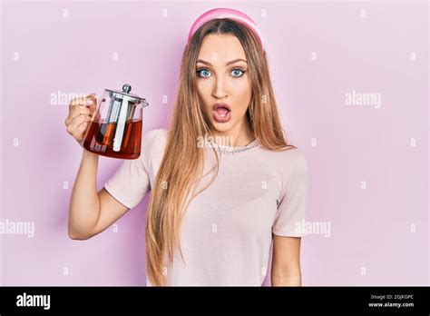 Jeune fille blonde tenant un pot de thé effrayée et stupéfait avec la bouche ouverte pour la
