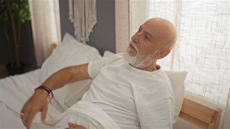 Mature Bald Man With A Beard Relaxing In A Cozy Indoor Bedroom Sitting On A Bed While Enjoying