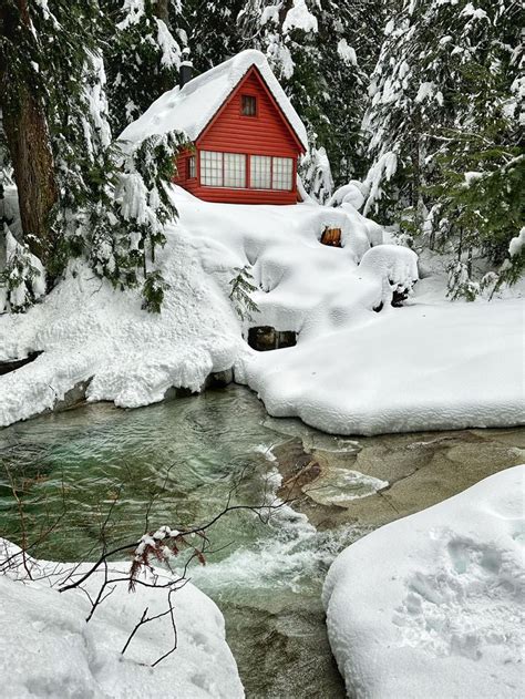 A Small Red Cabin In The Woods Covered In Snow Next To A Stream And Trees