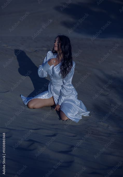 Glamorous Brunette Girl Poses On The Sand At Night In A Sexy Dress Showing Her Legs Stock Photo