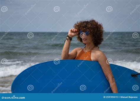 Attractive Mature Woman With Curly Hair Sunglasses And Bikini Posing Holding A Blue Surfboard