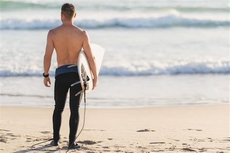 Premium Photo An Athletic Man Surfer With Naked Torso Standing By The Ocean Back View