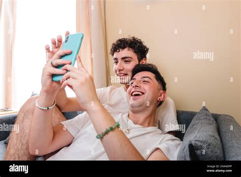 Snuggling Gay Couple In Pajama Laughing And Using The Mobile To Watch Entertaining Content Stock