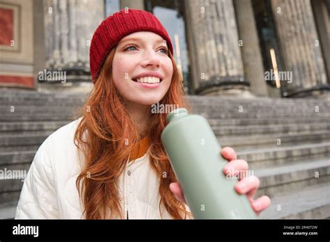 Portrait Of Redhead Woman Drinking From Thermos Sitting On Street Stairs And Enjoys Hot Drink