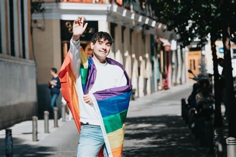Joven gay envuelto en una bandera del arco iris de pie saludando a la cámara en medio de la