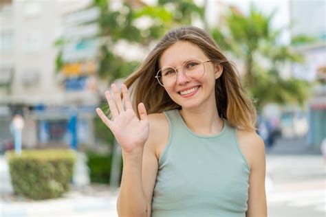 Junge blonde frau mit brille im freien salutiert mit der hand und glücklichem gesichtsausdruck