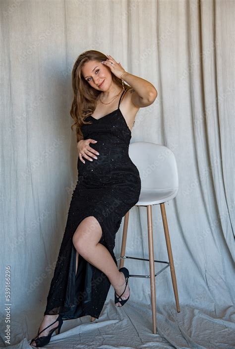 Beautiful Sexy Pregnant Brunette Woman In Black Dress Sitting On The Chair In Studio And Holding