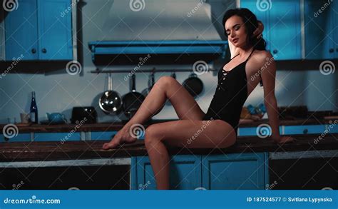 Girl In Black Bodysuit Lingerie Smiling Sitting Sensually On A Kitchen Island Table With One Leg